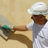 Muž v prilbe a ochranných okuliaroch aplikujúci žltú fasádnu omietku.