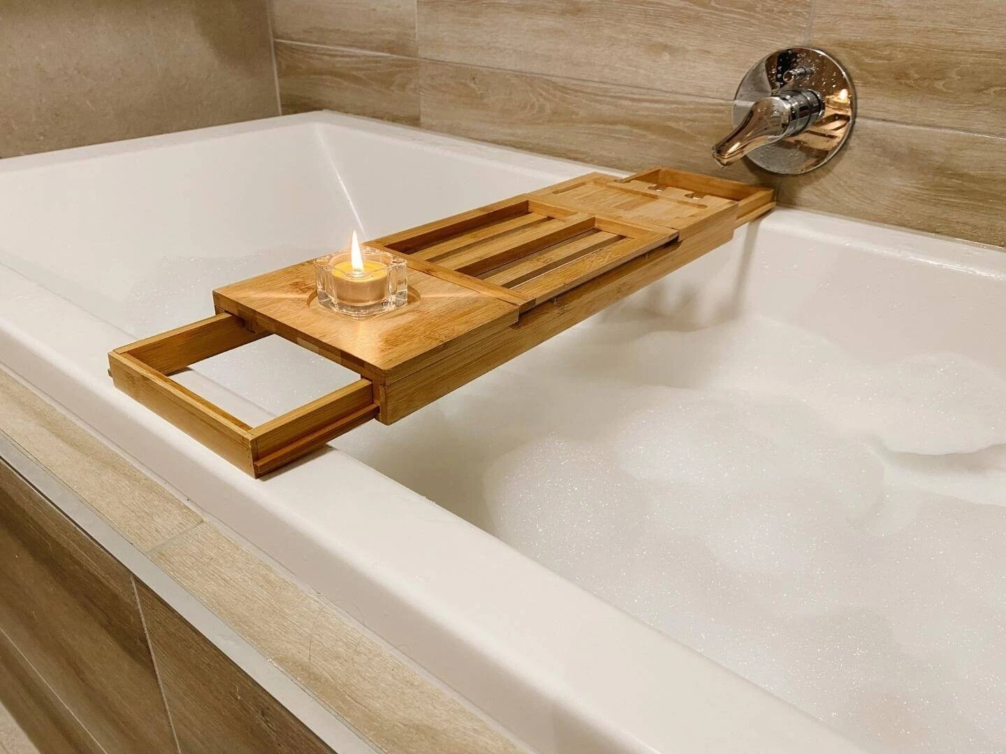 Zapálená medová sviečka na bambusovom stolčeku položenom na vani s penovým kúpeľom.