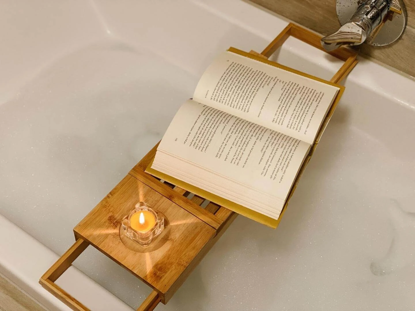 Otvorená kniha a zapálená sviečka položená na tácke na napustenej vani s penou.