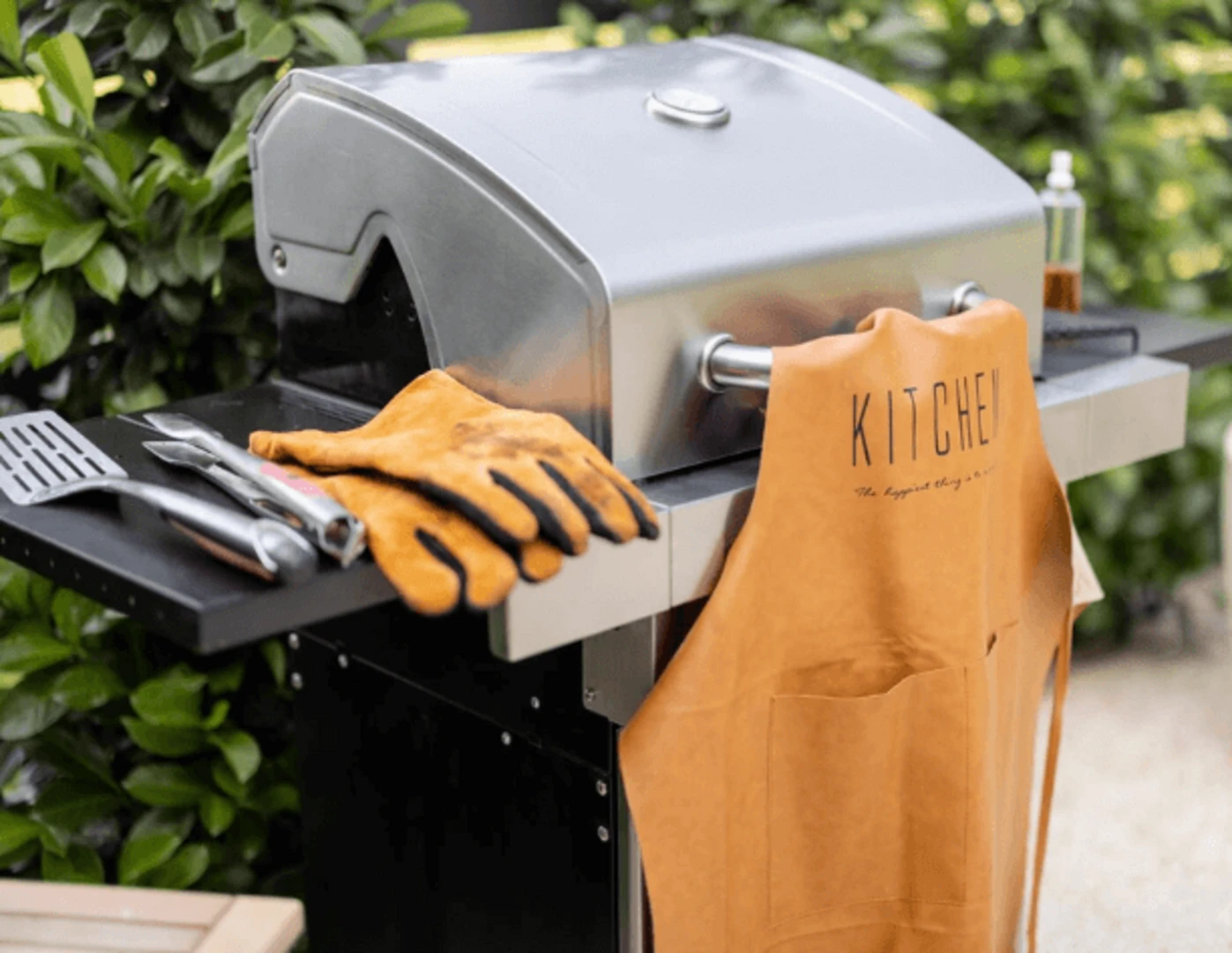 Lopatka, kliešte, rukavice a zástera položené na zatvorenom nerezovom grile.