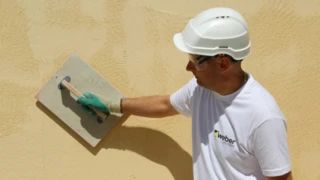 Muž v prilbe a ochranných okuliaroch aplikujúci žltú fasádnu omietku.